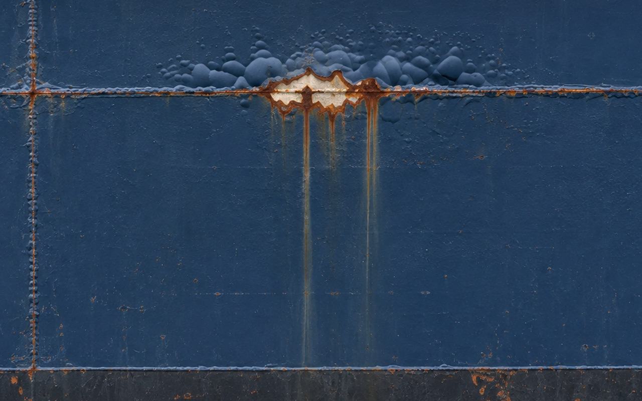 Close-up of a ship's hull: blistered coating and exposed primer along a horizontal plate seam, with rust streaks running down
